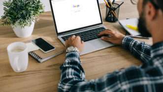 A man in a plaid shirt sits at a laptop open to the Google homepage. | Lightfieldstudiosprod | Dreamstime.com